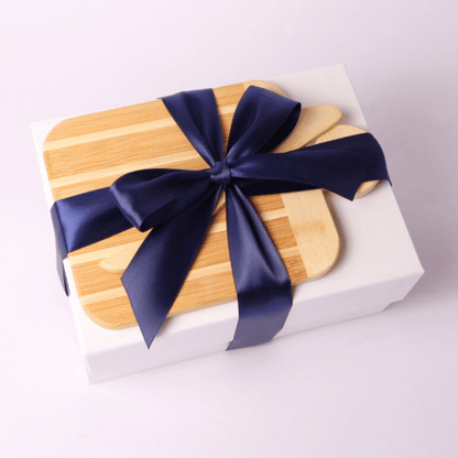 Simply Savory Gift Box wrapped with a navy blue satin bow and topped with a bamboo paddle board, perfect for hostess or housewarming gifts.