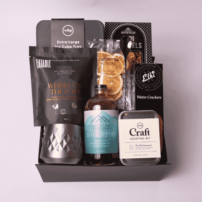 Sophisticated black gift box featuring bourbon-inspired treats: old fashioned cocktail syrup, whiskey tumbler, bar spoon, orange slices, pretzels, and crackers.